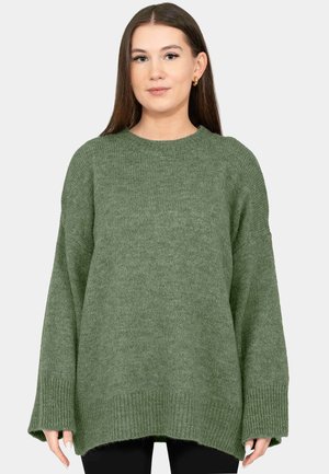 Junge Frau mit langen braunen Haaren, die einen lockeren, gerippten olivgrünen Pullover und goldene Creolen vor einem schlichten, hellen Hintergrund trägt.