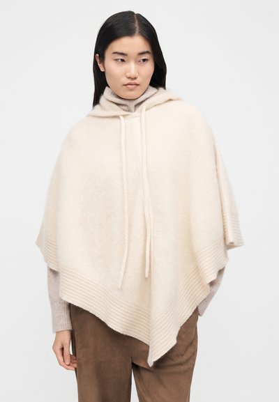 Poncho à capuche beige en tissu doux, avec un ourlet côtelé et des cordons de serrage, présentant une silhouette décontractée et carrée. Détail de surface texturée.