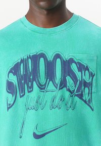 Zelena bombažna majica z velikim modrim grafičnim napisom "SWOOSH" in "just do it", dopolnjena z majhnim žepom na prsih.