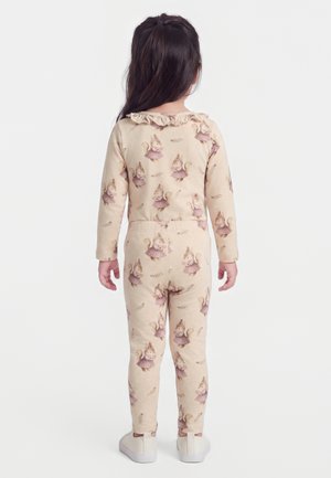 Jeune enfant aux longs cheveux foncés portant un pyjama beige avec des imprimés d'écureuils roses et des chaussures blanches à enfiler, debout de dos.