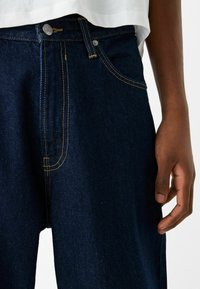 Tmavě modré džíny z denimu mají kontrastní šití, knoflíkový zapínání a přední kapsy. Viditelná textura zlepšuje vzhled látky.