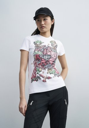 Γυναίκα που φοράει μαύρο καπέλο, λευκό μπλουζάκι με πολύχρωμο εκτύπωμα γης και πουλιού και μαύρο παντελόνι με τσερνά με φερμουάρ, ποζάροντας σε γκρι φόντο.