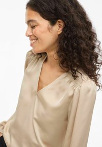 Beige satengbluse med V-hals, detaljer med rynkede ermer og glatt tekstur. Designet har en avslappet passform og subtil glans.