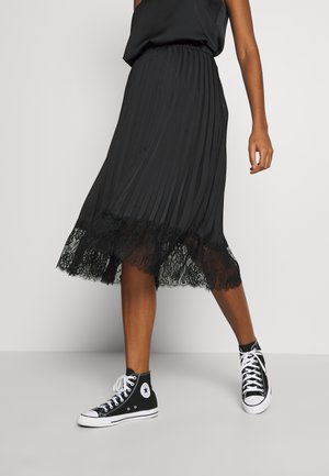 Vrouw draagt een zwarte geplooide midirok met kanten zoom en zwarte hoge sneakers, staand tegen een effen achtergrond.