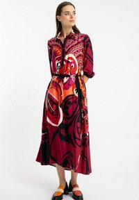 Kobieta w sukience z długim rękawem, zapinanej na guziki, z żywym, abstrakcyjnym wzorem w kolorze czerwonym, pomarańczowym i czarnym, połączona z czarnymi butami z pomarańczowymi i białymi paskami.