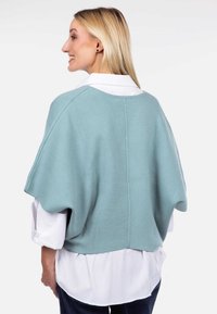 Jasnoniebieskie, dziergane ponczo z szerokimi rękawami i zaokrąglonym dekoltem, noszone na białej koszuli. Gładka tekstura, casualowy design.