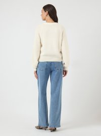 Kobieta stojąca tyłem, ubrana w białą bluzę, niebieskie szerokie spodnie dżinsowe i beżowe buty na koturnie, na tle jednolitego białego tła.