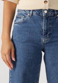 Nő világos bézs színű kötött kardigánban és közép derekú kék farmernadrágban, keze lazán a test mellett, egyszínű háttér előtt.