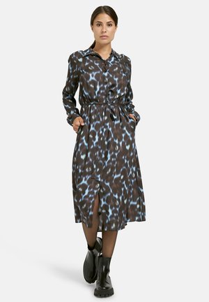 Frau steht mit den Händen in den Taschen, trägt ein langärmeliges, tailliertes, midi-kleid mit einem blau-braunen Muster und schwarze Knöchelstiefel.