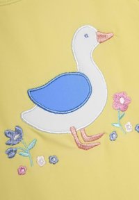 Una tela amarilla presenta un pato bordado en blanco con un ala azul y un pico coral, rodeado de diseños florales en colores pastel.