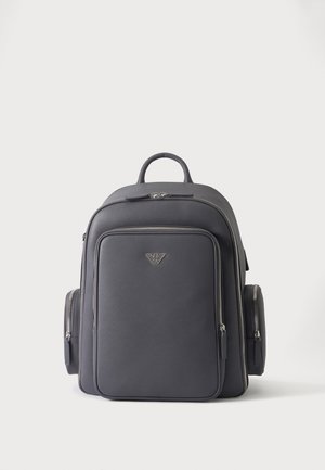 Sac à dos en cuir noir avec poches zippées à l’avant et sur les côtés, et un petit logo métallique au centre devant.