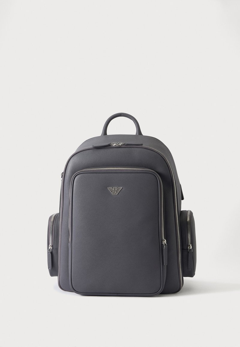 Sac à dos en cuir noir avec poches zippées à l’avant et sur les côtés, et un petit logo métallique au centre devant.