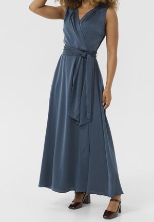 Kobieta ubrana w granatową sukienkę typu wrap bez rękawów z przewiązanym w talii paskiem oraz bordowe buty na słupku, stojąca na tle jednolitego tła.