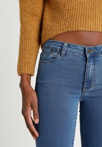 Ribbad senapsgul tröja med långa ärmar i kombination med högmidjade medelblå jeans som har framfickor och knappstängning.