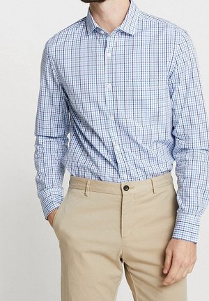 Homme portant une chemise à manches longues à carreaux bleus et blancs rentrée dans un pantalon beige, une main dans la poche.