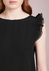 Blusa nera con scollatura tonda e decorazioni in ruffles trasparenti arricciati sulle maniche. Tessuto liscio e design aderente.