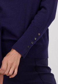 Marineblauwe gebreide trui met een geribde onderrand en knoopdetails op de manchetten. Materiaal lijkt zacht met een gladde textuur. Horizontale naad aanwezig.