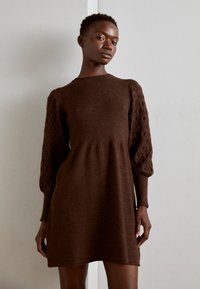 Ruskea neulemekko, jossa on ribatty yläosa ja teksturoitu, puffi hiha. Mekossa on levenevä siluetti ja pyöreä pääntie.
