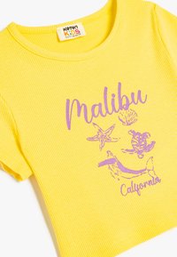 Camiseta de algodón amarilla con rayas, que presenta texto impreso en morado "Malibu" y "California" junto con ilustraciones temáticas del mar como una estrella de mar y una ballena.