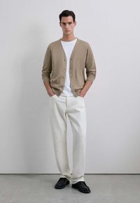 Cárdigan beige con botones, usado sobre una camiseta blanca. Pantalones blancos de corte holgado y zapatos negros completan el conjunto. Diseño minimalista.