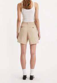 Pantalones cortos beige con un corte relajado, dos bolsillos traseros con botones y una tela suave y ligera. Llevado con una camiseta sin mangas blanca y zapatos negros.