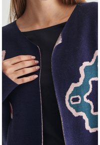 Námornícký pletený kardigan s geometrickým vzorem v teal a růžové barvě, s předním zipem a dlouhými rukávy. Měkká textura a volný střih.