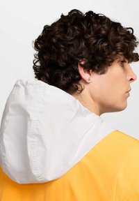 Chubasquero amarillo con capucha blanca. Material ligero, corte relajado, costuras visibles. La modelo tiene el cabello rizado y un pequeño pendiente.