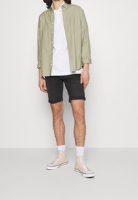 Chemise boutonnière vert clair sur un t-shirt blanc, shorts noirs retroussés, et baskets montantes beiges avec semelles blanches.