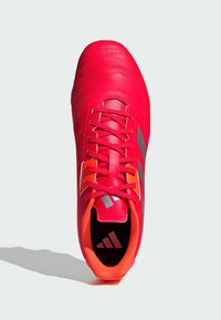 Zapatilla de fútbol roja con parte superior de cuero texturizado, seis ojetes, acentos naranjas y un logo negro y plateado en la lengüeta y el costado.