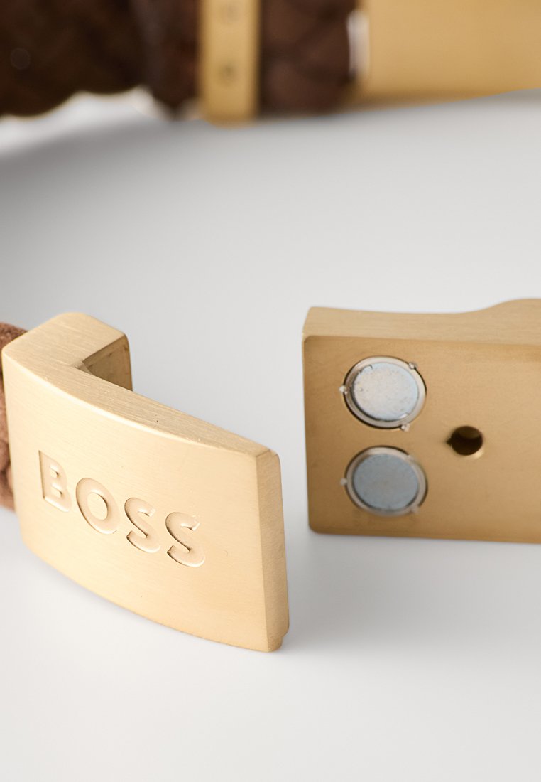 Ceinture en cuir tressé marron avec une boucle en métal doré rectangulaire portant le mot "BOSS" gravé dessus. Fini lisse, fermeture magnétique.