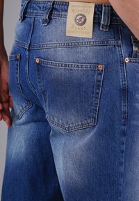 Blue denim jeans with a faded texture, featuring a square label at the waistband, copper rivets, and two back pockets.