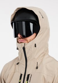 Giacca impermeabile beige con chiusura a zip, cappuccio regolabile e fori di ventilazione. Indossata con un casco nero e occhiali protettivi neri oversize.