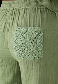 Pantalones de tela verde claro y texturizada que presentan un bolsillo de estilo crochet con un patrón geométrico en la parte trasera. Tienen un ajuste suave y relajado.