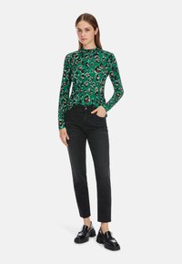 Grünes Langarmoberteil mit Leopardenmuster, kombiniert mit schwarzen Jeans. Das Model trägt glänzende schwarze Plateauslipper. Einfache Hintergrund.