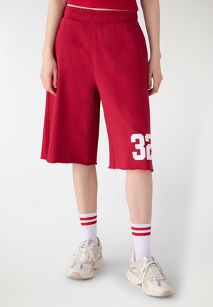 Persona che indossa pantaloncini rossi al ginocchio con il numero 32 sulla gamba sinistra, scarpe da ginnastica bianche e calzini bianchi con strisce rosse.