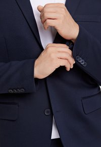 Blazer azul marino con una textura suave, cierre de un botón y detalle de puño con cuatro botones. Presenta un cuello con muesca y bolsillos laterales.