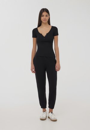 Top corto nero a maniche corte con scollatura a V e dettaglio di bottoni, abbinato a pantaloni jogger neri con polsini a coste, indossato con sneakers bianche.