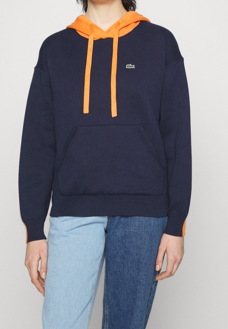 Personne portant un sweat à capuche bleu marine avec une capuche orange et des cordons, une poche kangourou, et un jean bicolore bleu, clair à gauche et foncé à droite.