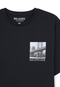 Czarny bawełniany t-shirt o luźnym kroju, z małą czarno-białą grafiką mostu oraz tekstem na lewej części klatki piersiowej.