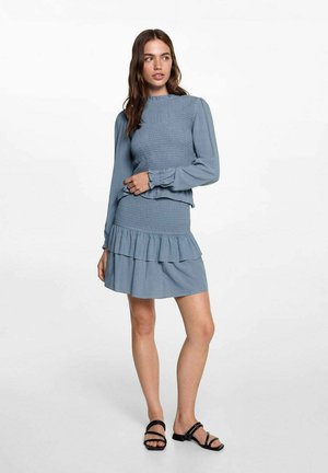 Ensemble deux pièces bleu clair comprenant un haut à manches longues smocké et une jupe à volants superposés. Tissu texturé et coupe ample. Sandales noires.