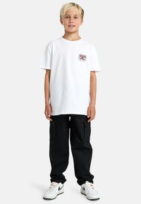 T-shirt bianca in cotone con una stampa sul petto, abbinata a pantaloni cargo neri e sneakers bianche con dettagli verdi.