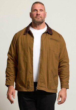 Homme aux cheveux courts et à la barbe, portant une veste marron zippée par-dessus une chemise blanche, debout devant un fond clair uni.