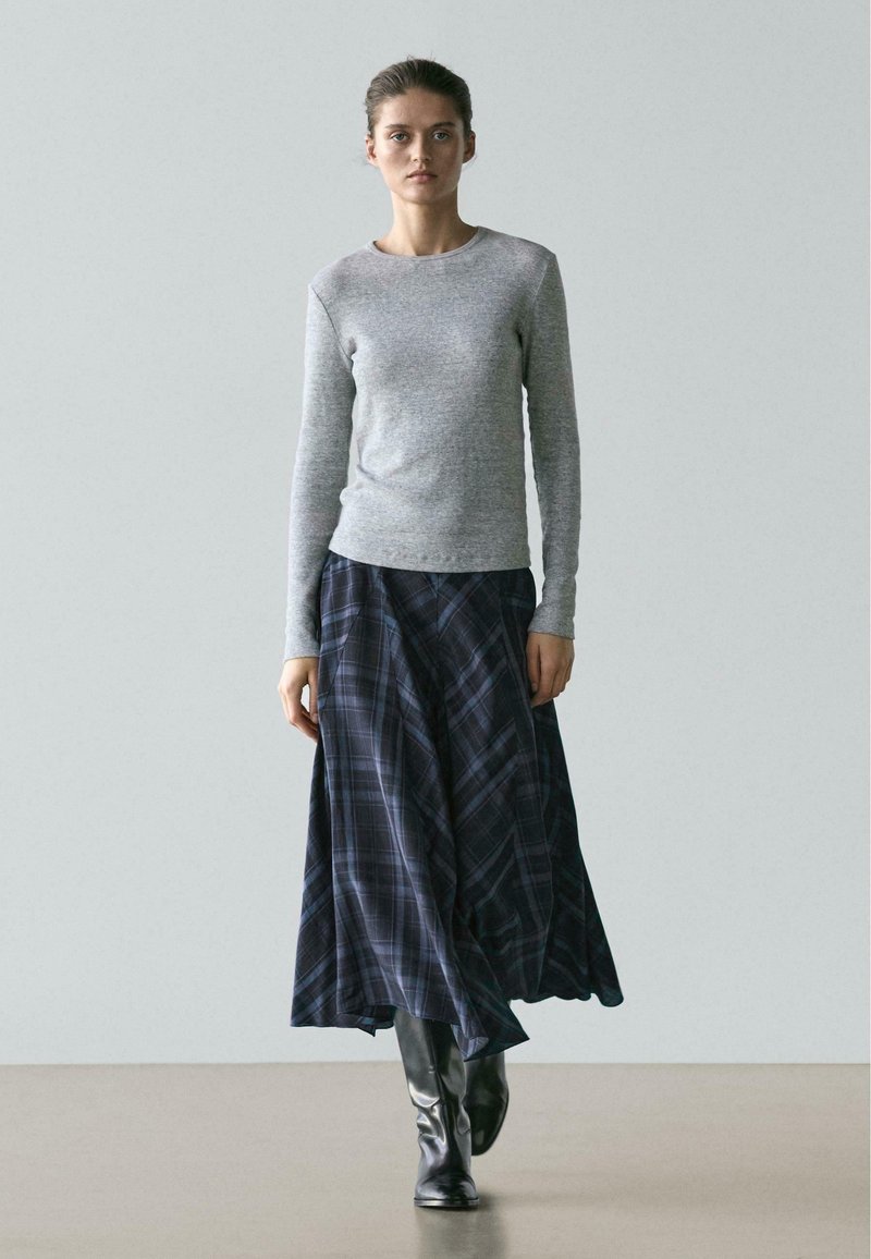 Kobieta ubrana w szarą koszulę z długim rękawem, zwiewną ciemną spódnicę w kratę oraz czarne kozaki za kolano, stojąca na neutralnej podłodze i tle.