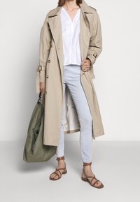 Trench beige con grandes botones, combinado con una camisa blanca, jeans azul claro y sandalias marrón con tiras. Lleva una bolsa de tela verde.