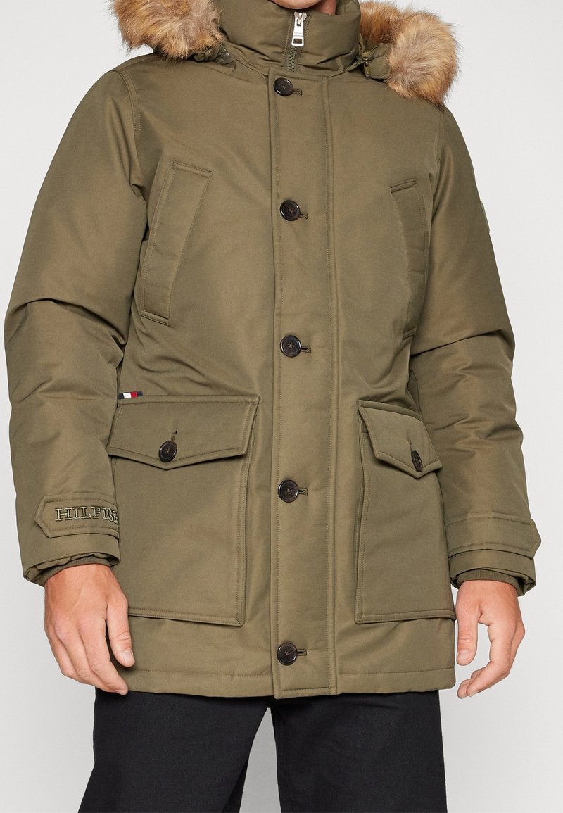 Olivengrøn parka med faux fur hættekant, knaplukninger og flere frontlommer. Har logodetalje på ærmet. Glat overflade.