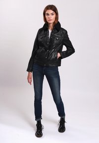 Schwarze Lederjacke mit Knopfverschluss, Brusttaschen und strukturiertem Kragen, getragen über einem weißen Hemd und kombiniert mit dunklen Jeans und Stiefeln.