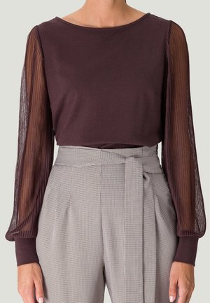 Woman wearing a dark purple top with sheer pleated long sleeves and high-waisted gray checkered pants with a tied belt.