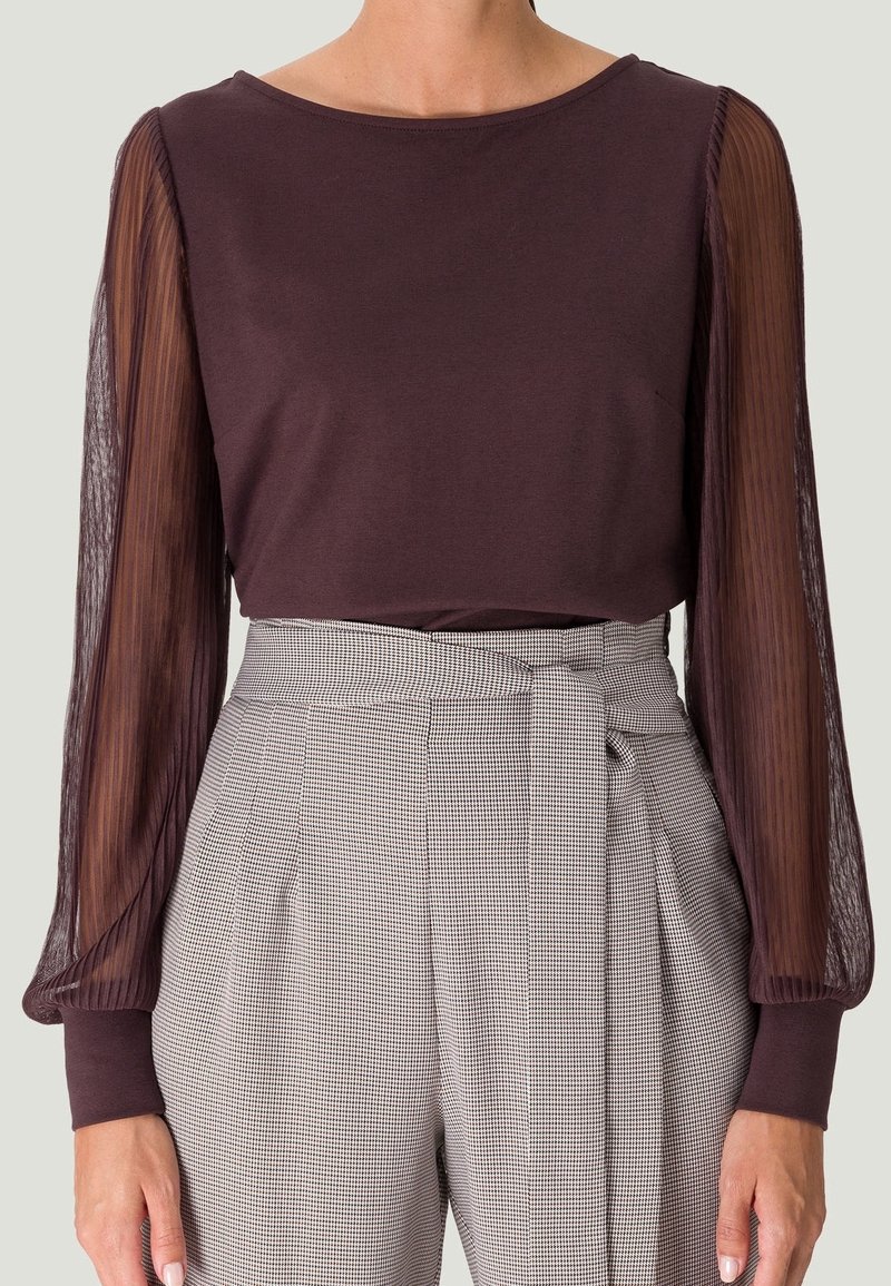 Femme portant un haut violet foncé avec des manches longues plissées transparentes et un pantalon taille haute gris à carreaux avec une ceinture nouée.