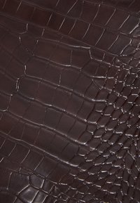 Ruskea krokotiilin kuvioitu nahka, jossa on teksturoitu pinta, erottuvat suomut ja kiiltävä viimeistely. Huomattavat kuviot luovat visuaalista syvyyttä.