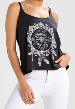 Vrouw draagt een zwarte tanktop met een wit geometrisch mandala- en veerontwerp, gecombineerd met een witte broek, hand rust op heup.